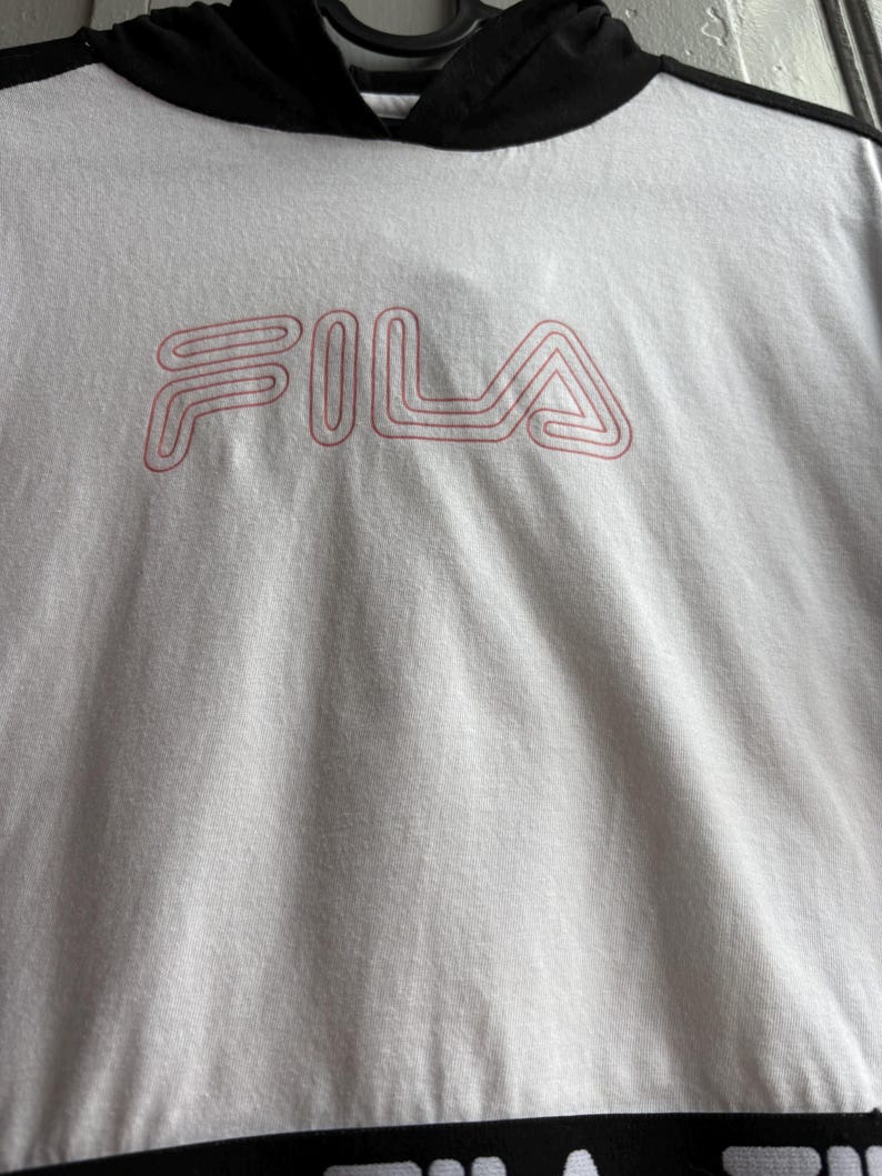 May include: White hooded sweatshirt with the brand name "FILA" in pink lettering. The sweatshirt has a black hood and a black band at the bottom. The fabric appears to be a soft, textured material.