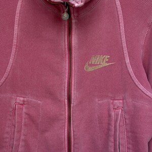 K&ouml;nnte beinhalten: Staubrosa Nike-Jacke mit Rei&szlig;verschluss. Die Jacke hat einen durchgehenden Rei&szlig;verschluss, zwei Vordertaschen und das goldene Nike-Logo auf der Brust. Der Stoff hat eine strukturierte Optik.