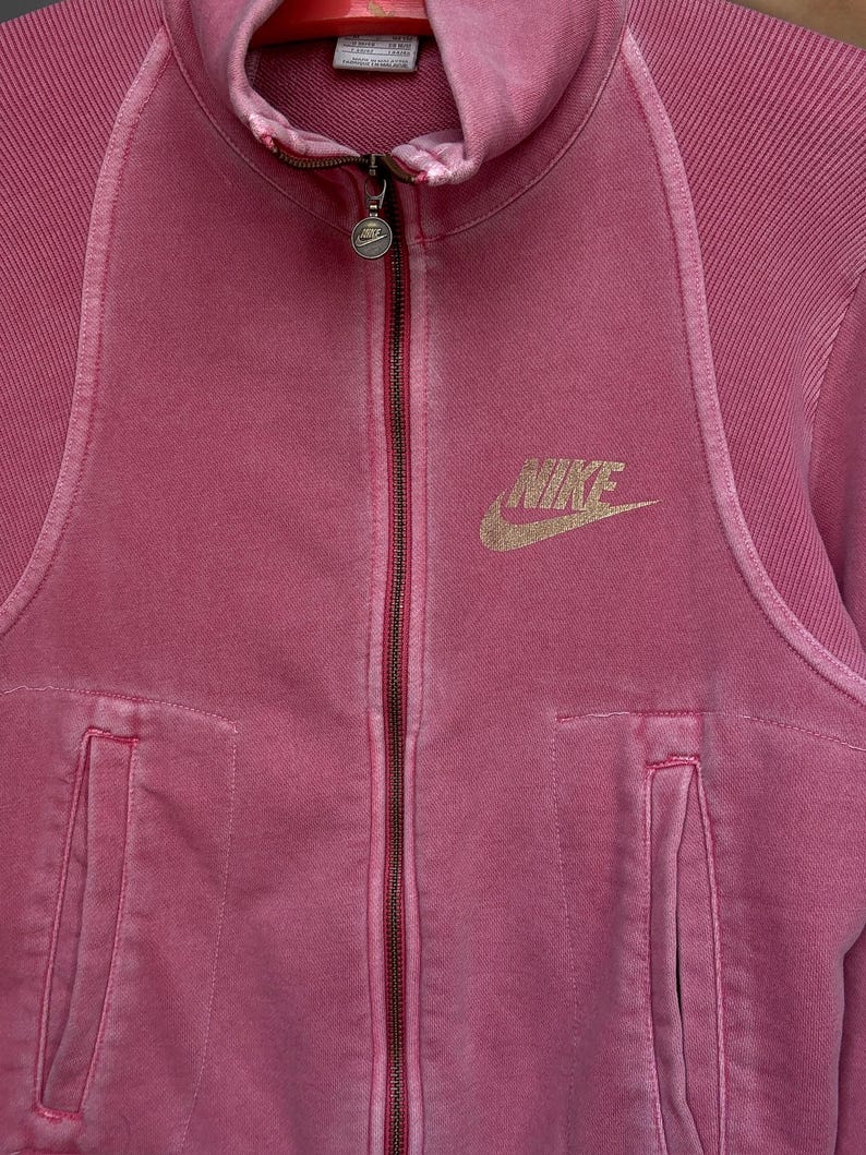 K&ouml;nnte beinhalten: Staubrosa Nike-Jacke mit Rei&szlig;verschluss. Die Jacke hat einen durchgehenden Rei&szlig;verschluss, zwei Vordertaschen und das goldene Nike-Logo auf der Brust. Der Stoff hat eine strukturierte Optik.