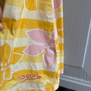 May include: Yellow and white striped fabric with large pink and white floral designs. The fabric has the word "ripcurl" embroidered in pink. The fabric is folded, showing the edge of the material.