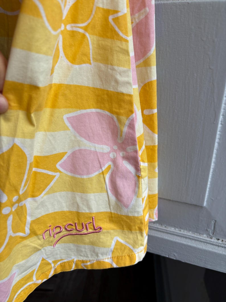 May include: Yellow and white striped fabric with large pink and white floral designs. The fabric has the word "ripcurl" embroidered in pink. The fabric is folded, showing the edge of the material.