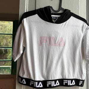 May include: White crop top with a black hood and sleeves, featuring the pink "FILA" logo on the front. The bottom band has the "FILA" logo in white on a black background. The top is hanging on a black hanger.