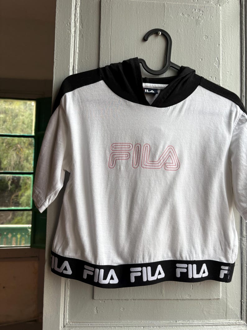 May include: White crop top with a black hood and sleeves, featuring the pink "FILA" logo on the front. The bottom band has the "FILA" logo in white on a black background. The top is hanging on a black hanger.