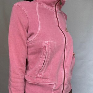 K&ouml;nnte beinhalten: Eine rosa Jacke mit Rei&szlig;verschluss und hohem Kragen. Die Jacke hat einen gerippten Einsatz an der Schulter und eine Tasche auf der Vorderseite. Der Rei&szlig;verschluss ist in einem dunkleren Rosaton gehalten. Die Jacke hat ein kleines Logo auf der Brust.