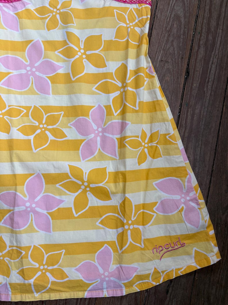 May include: A yellow and white striped dress with a floral pattern of pink and yellow flowers. The dress has a pink trim at the top and the Rip Curl logo near the bottom.