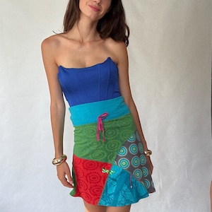 May include: A strapless blue top and patchwork mini skirt. The skirt features panels of green, red, and teal fabrics with various patterns, including circles and floral designs. A pink drawstring cinches the waist. Gold bracelets adorn the wrists.