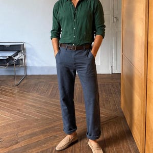Peut inclure: Une personne porte une chemise vert foncé, un pantalon bleu marine, une ceinture marron et des mocassins en daim beige. Le pantalon est retroussé aux chevilles. La tenue est décontractée et élégante.