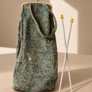 Può includere: Una pochette in tessuto verde con motivo paisley e chiusura a scatto in metallo dorato, dotata di un manico in tessuto coordinato. Accanto alla pochette si trovano due ferri da maglia grigi con punte gialle, contrassegnati dal numero 6 in blu.
