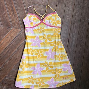 May include: A yellow, white, and pink floral print dress with spaghetti straps. The dress features a striped pattern with pink flowers and a pink trim. The Rip Curl logo is visible on the bottom.