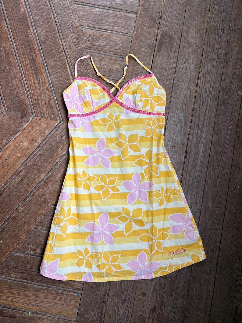 May include: A yellow, white, and pink floral print dress with spaghetti straps. The dress features a striped pattern with pink flowers and a pink trim. The Rip Curl logo is visible on the bottom.