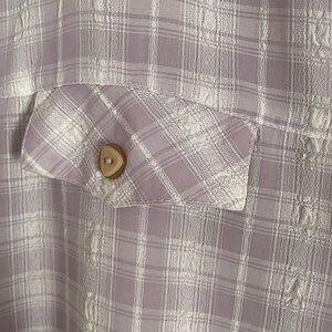 May include: A close-up of a light purple and white plaid fabric with a small, round, brown button. The fabric has a subtle embossed pattern.
