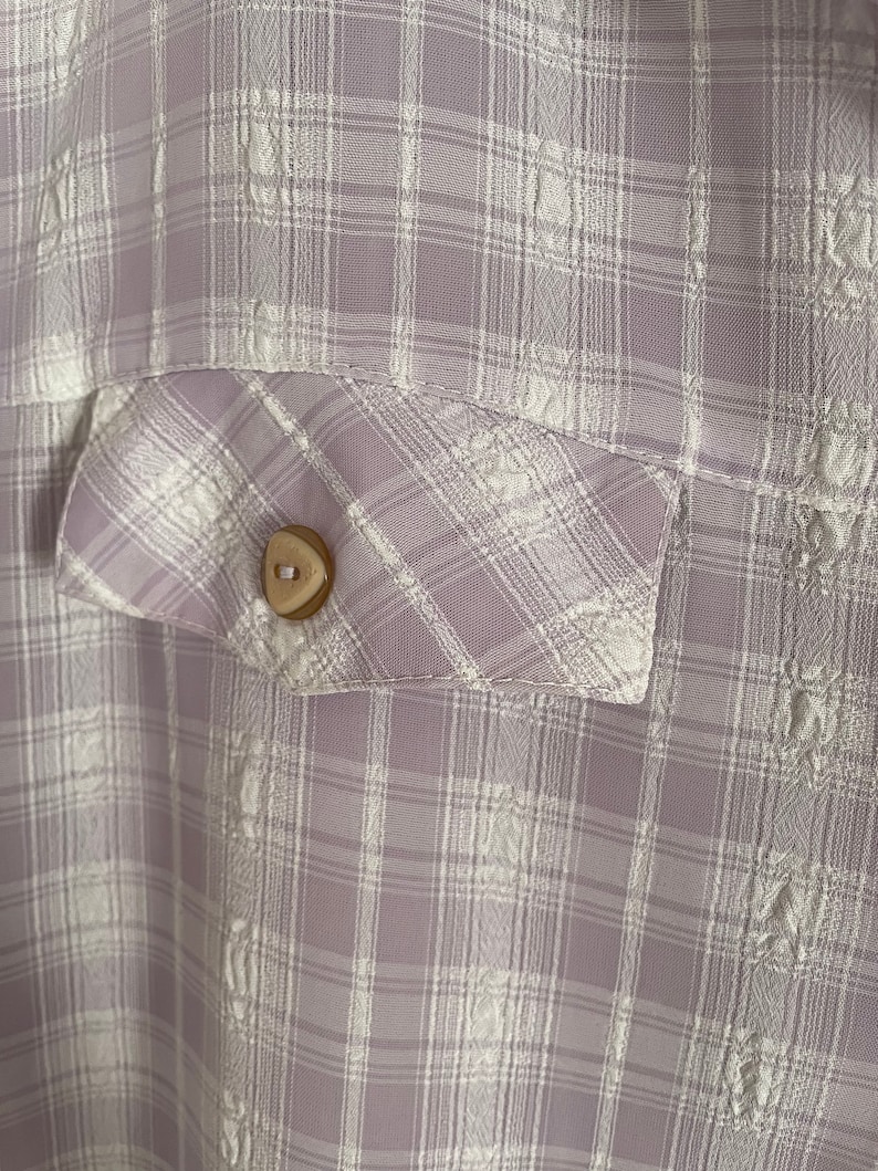 May include: A close-up of a light purple and white plaid fabric with a small, round, brown button. The fabric has a subtle embossed pattern.
