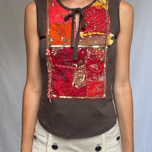 Op de afbeelding: Een mouwloze bruine top met een patchwork ontwerp in tinten rood, oranje en geel. Het patchwork is versierd met pailletten en een strik bij de halslijn. De top wordt gecombineerd met een lichtgekleurde broek.