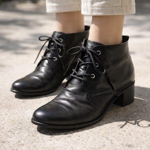 Könnte beinhalten: Schwarze Leder-Ankle Boots mit Schnürdesign und kleinen Blockabsätzen. Die Stiefel haben eine runde Spitze und werden mit hellen Hosen getragen. Die Stiefel sind aus glattem, glänzendem Leder gefertigt.