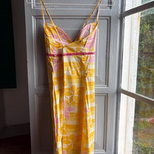 May include: A yellow sundress with a floral pattern and thin straps hangs on a hanger. The dress features a V-neckline with pink lining and a pink trim at the waist. The floral design is in pink and white.
