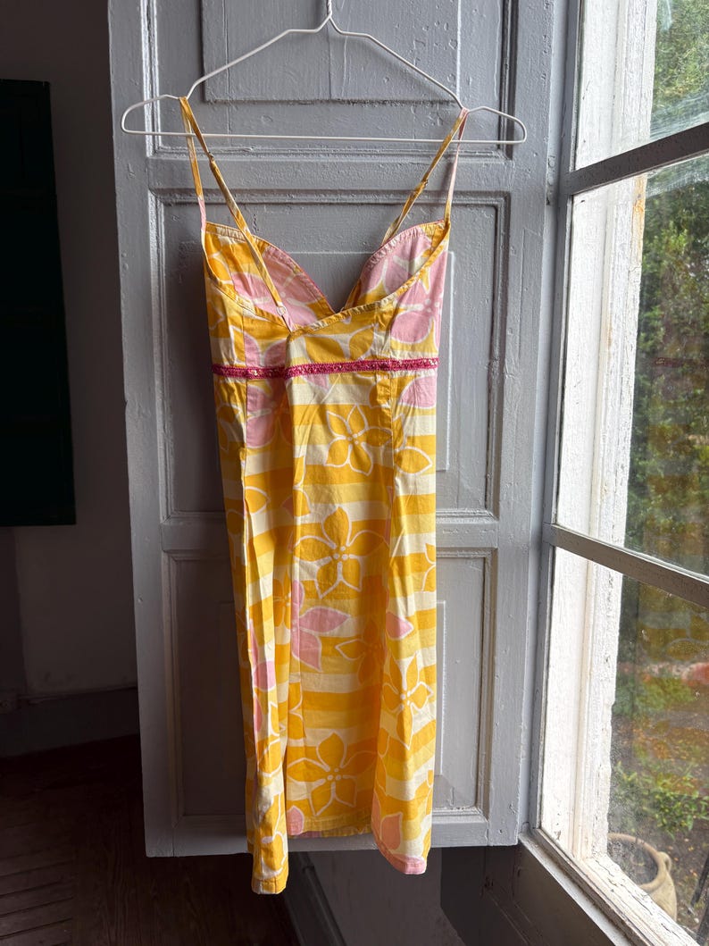May include: A yellow sundress with a floral pattern and thin straps hangs on a hanger. The dress features a V-neckline with pink lining and a pink trim at the waist. The floral design is in pink and white.