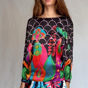 May include: A long-sleeved blouse with a vibrant, colourful floral pattern. The top features a black background with a pink scalloped design and a bold, abstract floral print in shades of pink, green, orange, and blue. The sleeves are long and the neckline is a scoop neck.
