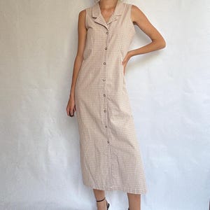 May include: A sleeveless, button-down dress in a beige and white plaid pattern. The dress has a collared neckline and falls to mid-calf. The dress features small, round buttons down the front. The dress is made of a lightweight fabric.