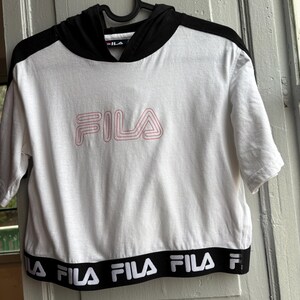 May include: White short-sleeved crop top with a black hood and black trim. The word "FILA" is printed in pink on the front. The bottom hem features the word "FILA" in white on a black band. The top is hanging on a black hanger.