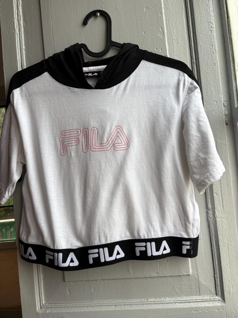 May include: White short-sleeved crop top with a black hood and black trim. The word "FILA" is printed in pink on the front. The bottom hem features the word "FILA" in white on a black band. The top is hanging on a black hanger.
