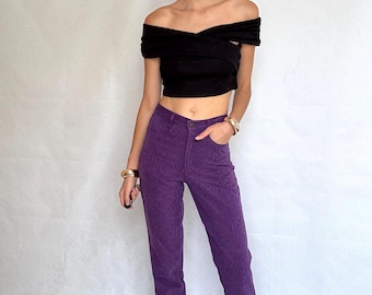 Könnte beinhalten: Ein schwarzes schulterfreies Oberteil kombiniert mit einer hoch taillierten, geraden Hose in Lila. Die Hose hat ein subtiles strukturiertes Muster. Das Model trägt goldene Armbänder und weiße Absätze. Das Outfit ist vor einem schlichten weißen Hintergrund.