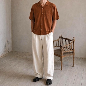 May include: A person wearing a short-sleeved, button-down shirt in a warm brown color, paired with wide-leg, off-white pants. The outfit is completed with black shoes. A wooden chair is in the background.