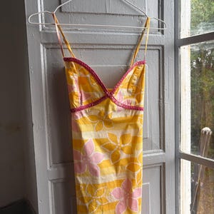 May include: A yellow sundress with pink floral and striped patterns, hanging on a white hanger. The dress has thin straps and pink trim along the neckline. The dress is made of a lightweight fabric.