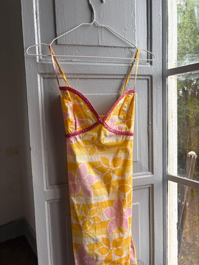 May include: A yellow sundress with pink floral and striped patterns, hanging on a white hanger. The dress has thin straps and pink trim along the neckline. The dress is made of a lightweight fabric.