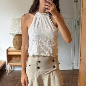 Può includere: Un top bianco senza maniche con scollo all'americana, intricato ricamo inglese e orlo smerlato, abbinato a una minigonna in maglia beige a vita alta, con un pannello frontale strutturato e quattro bottoni circolari scuri su due file.