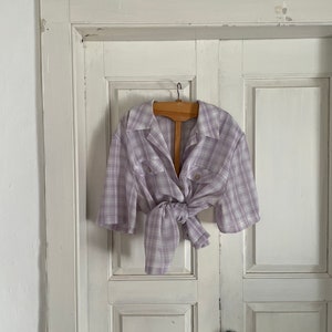 May include: A light purple and white plaid shirt with short sleeves, tied at the waist, hanging on a wooden hanger against a white door.
