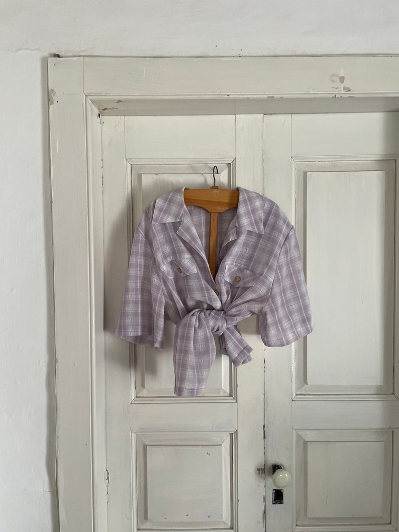 May include: A light purple and white plaid shirt with short sleeves, tied at the waist, hanging on a wooden hanger against a white door.
