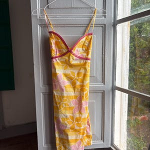 May include: Yellow sundress with pink and white floral pattern and horizontal stripes. The dress has thin straps and a pink trim along the neckline. The dress is hanging on a white hanger.