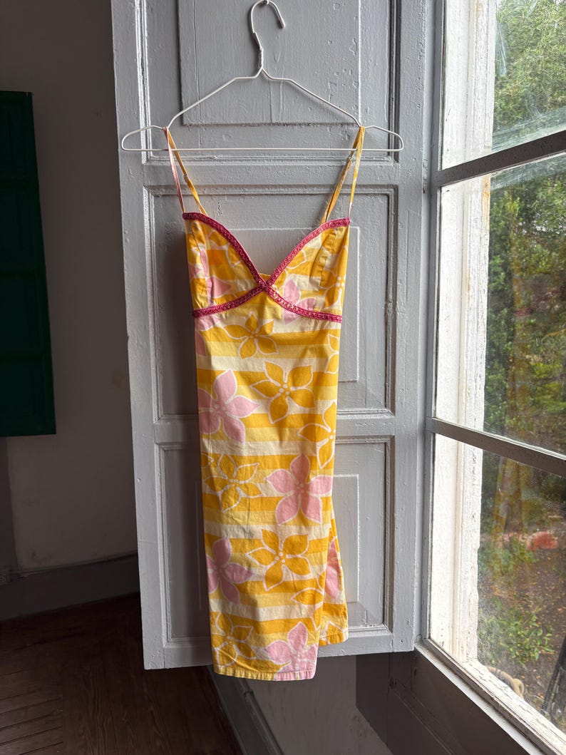 May include: Yellow sundress with pink and white floral pattern and horizontal stripes. The dress has thin straps and a pink trim along the neckline. The dress is hanging on a white hanger.