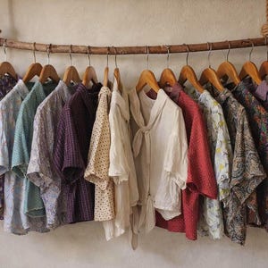 May include: A selection of patterned shirts hanging on wooden hangers from a natural branch. The shirts feature a variety of colours and patterns, including floral, polka dot, and striped designs. The shirts are made of lightweight fabric.