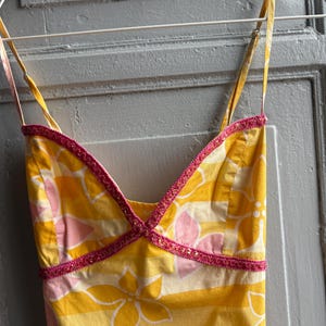 May include: A yellow sundress with a floral pattern in white and pink, hanging on a white hanger. The dress has thin straps and a V-neckline trimmed with pink beading. The fabric has horizontal stripes.
