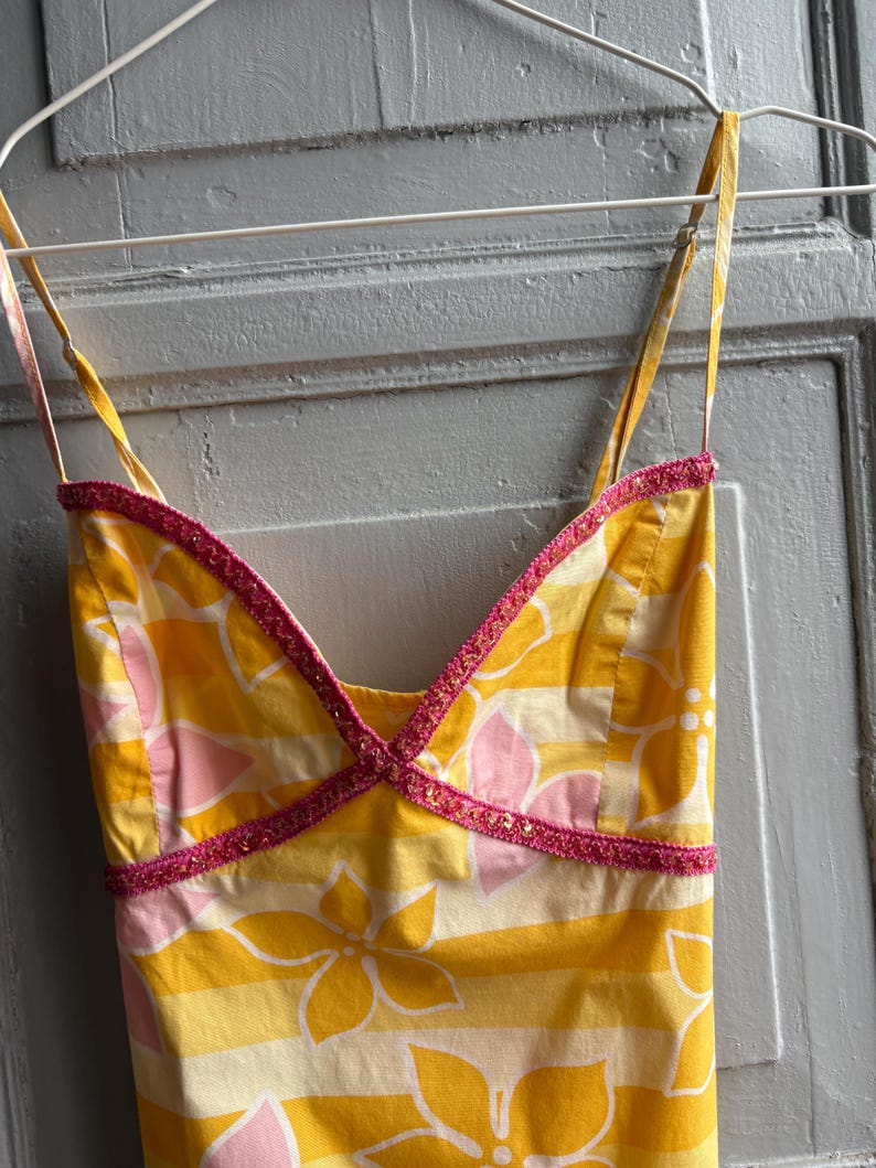 May include: A yellow sundress with a floral pattern in white and pink, hanging on a white hanger. The dress has thin straps and a V-neckline trimmed with pink beading. The fabric has horizontal stripes.
