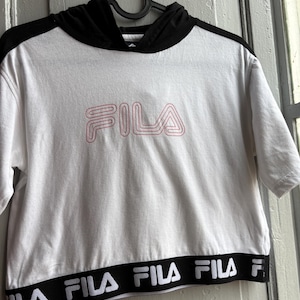 May include: White short-sleeved hoodie with a black hood and trim. The word "FILA" is displayed in pink lettering on the front. The bottom hem features a black band with the brand name in white.