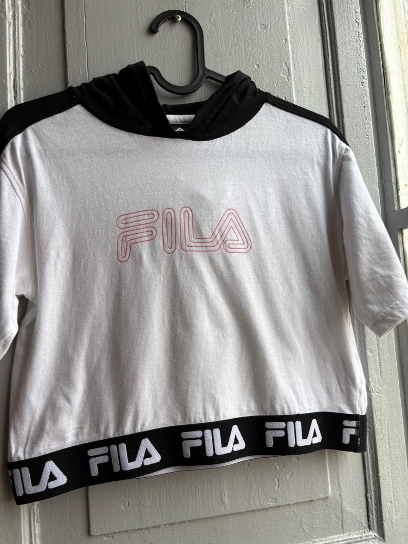 May include: White short-sleeved hoodie with a black hood and trim. The word "FILA" is displayed in pink lettering on the front. The bottom hem features a black band with the brand name in white.