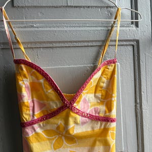May include: A yellow sundress with a floral pattern and thin straps hangs on a white hanger. The dress features a pink and yellow floral design with horizontal stripes. The neckline and straps are trimmed with pink beading.