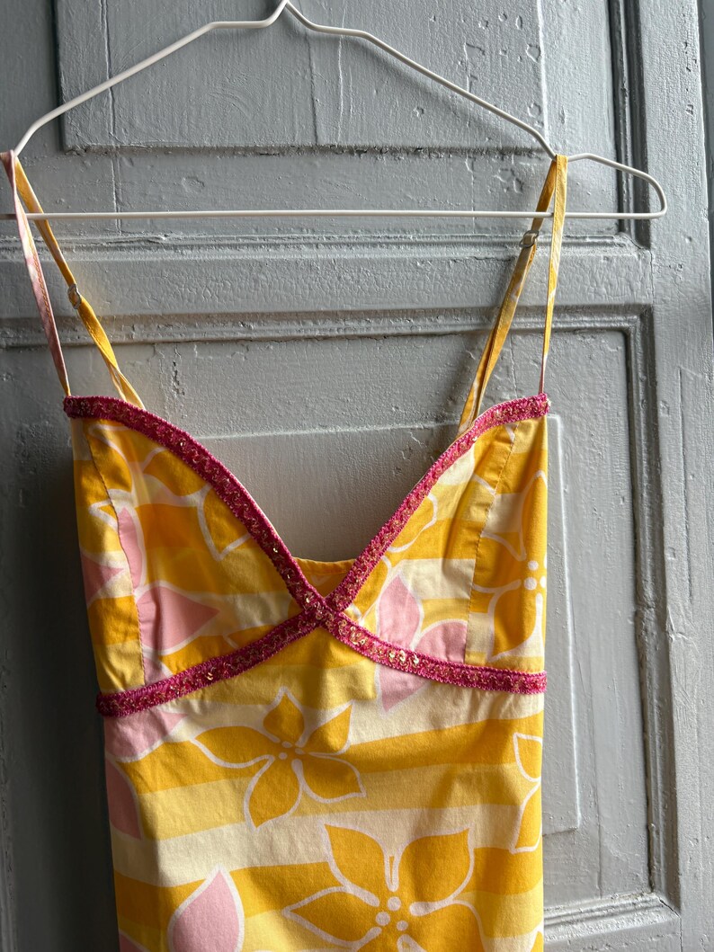 May include: A yellow sundress with a floral pattern and thin straps hangs on a white hanger. The dress features a pink and yellow floral design with horizontal stripes. The neckline and straps are trimmed with pink beading.