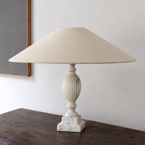 May include: A table lamp featuring a white marble base with gray veining and a fluted, urn-shaped central column. The lamp is topped with a wide, conical lampshade made of a textured, light beige fabric that provides a neutral, clean aesthetic.