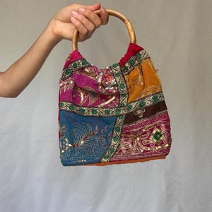 May include: A patchwork handbag with a wooden handle. The bag features a variety of colorful fabrics, including pink, blue, yellow, and brown, with intricate embroidery and sequin details. The handle is a natural wood color.