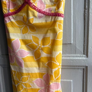 May include: Yellow and white striped sundress with large pink and yellow floral pattern. The dress has a V-neckline trimmed with pink sequins. The fabric is a lightweight cotton, perfect for warm weather.