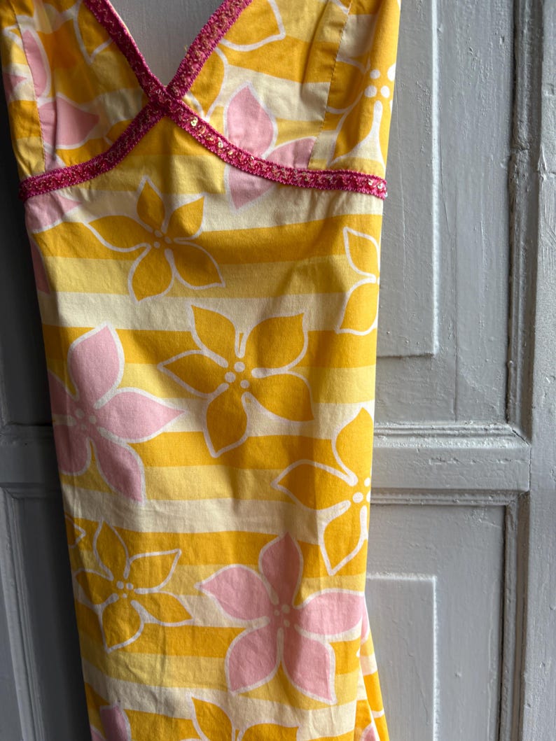 May include: Yellow and white striped sundress with large pink and yellow floral pattern. The dress has a V-neckline trimmed with pink sequins. The fabric is a lightweight cotton, perfect for warm weather.