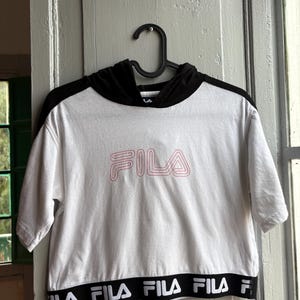May include: White crop top with a black hood and short sleeves, hanging on a black hanger. The word "FILA" is printed in pink on the front. The bottom hem features the brand name in a repeating black and white pattern.