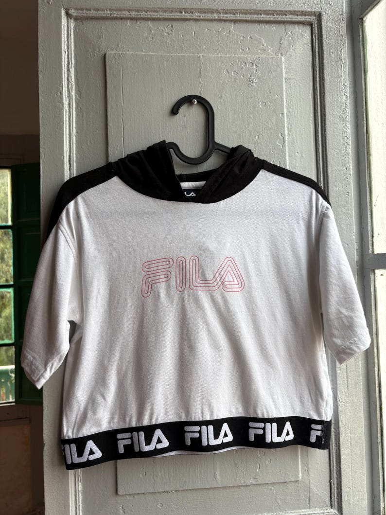 May include: White crop top with a black hood and short sleeves, hanging on a black hanger. The word "FILA" is printed in pink on the front. The bottom hem features the brand name in a repeating black and white pattern.