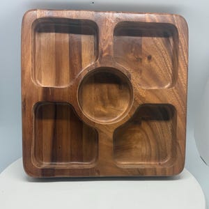 Puede incluir: Una bandeja de servir cuadrada de madera con un compartimento central redondo y cuatro compartimentos rectangulares. La bandeja está hecha de madera marrón claro con patrones de vetas visibles, ideal para servir aperitivos.
