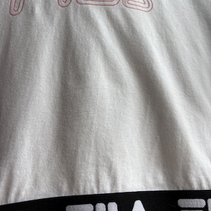 May include: Close-up of a white t-shirt with the brand name "FILA" in red, outlined letters at the top. A black band with the brand name "FILA" in white letters is at the bottom of the shirt. The fabric has visible texture.