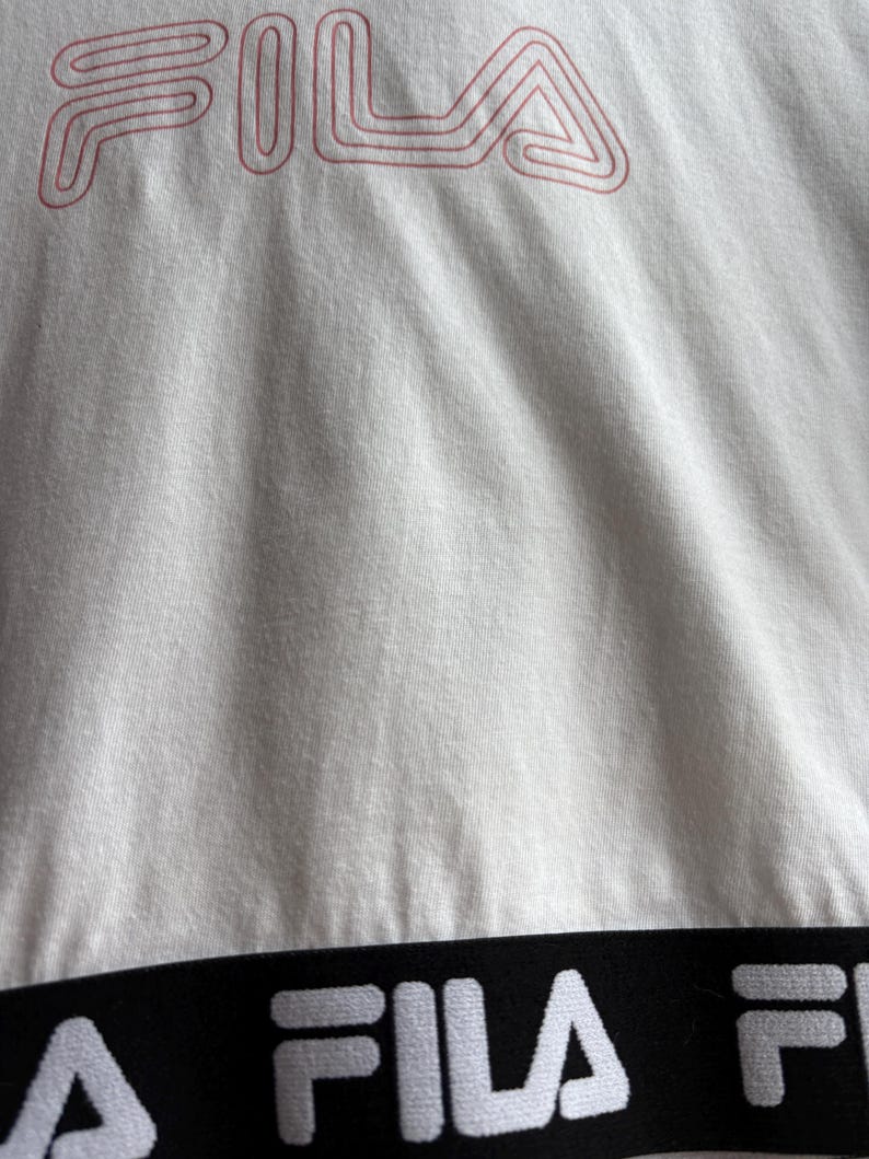 May include: Close-up of a white t-shirt with the brand name "FILA" in red, outlined letters at the top. A black band with the brand name "FILA" in white letters is at the bottom of the shirt. The fabric has visible texture.