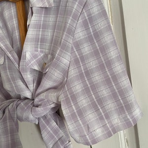 May include: A light purple and white plaid shirt with short sleeves and a button-up collar. The shirt is tied at the waist.
